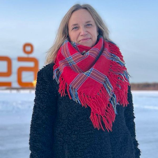 En kvinna står utomhus framför en stor skylt som visar texten ”Luleå”. Hon bär en svart jacka och en röd- och blåmönstrad halsduk. I bakgrunden syns vinterklädda människor och ett snöbelagt landskap.