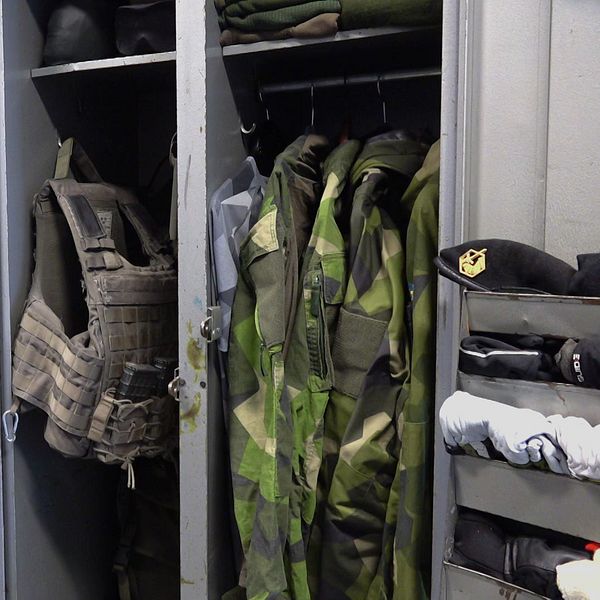 På vänster sida visas en person i uniform, medan höger sida visar en militär garderob fylld med olika typer av uniformsplagg och skyddsutrustning. Garderoben innehåller kamouflagekläder och en skottsäker väst, vilket ger en inblick i den militära utrustningen. Försvarsmakten, kvinnor i försvaret, Skövde, P4 Skövde.