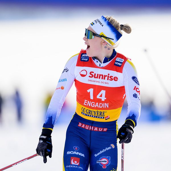 En kvinna deltar i en längdskidåkningstävling iförd nummer 14. Hon har en dräkt med sponsorloggor och håller i skidstavar, omgiven av ett snöklätt landskap. Bakgrunden är oskarp med flera människor synliga på avstånd.