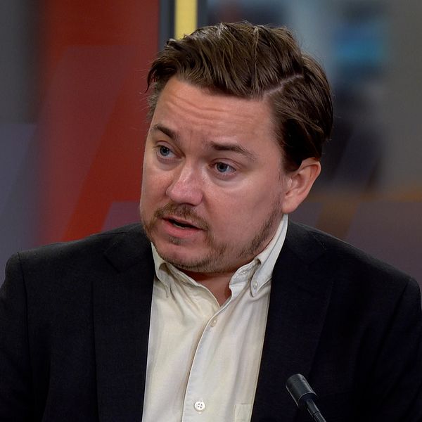 Bilden är en delad bild som visar två personer i olika miljöer. Den vänstra sektionen visar en person i militäruniform som står bakom en scen med en skylt med texten ”Folk och Försvar”. Den högra sektionen visar en person i en mörk kostym som sitter i en tv-studio.