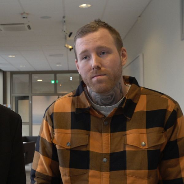 Två män står i en kontorsmiljö med moderna fönster i bakgrunden. Den första personen bär en mörk kavaj och en ljus skjorta, medan den andra personen har på sig en rutmönstrad skjorta i orange och svart. De ser mot kameran och verkar vara involverade i en diskussion eller presentation. De två männen är Arvikas kommundirektör Hans Karlsson och Robin Dahlén.