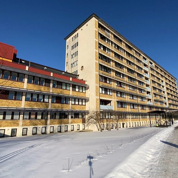 En delad bild med två sektioner. Den vänstra sektionen visar en person vid ett podium med en dator där namnskylten indikerar ”Maria Strandberg”. Den högra sektionen visar en utomhusbild av ett stort, flervåningshus beläget i ett vinterlandskap.