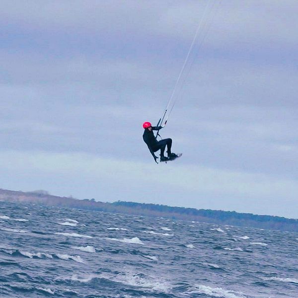 En delad bild som visar två scener relaterade till kitesurfing. Den vänstra sidan visar en person som håller en kitesurfingutrustning med en färgglad kite i bakgrunden. Den högra sidan visar en kitesurfare som svävar över vattnet, iförd en röd hjälm och svart dräkt, i en vindig miljö.