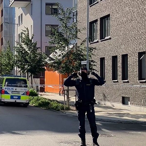 En polis står framför avspärrningar i ett bostadsområde med flerfamiljshus. Polisen håller för öronen.