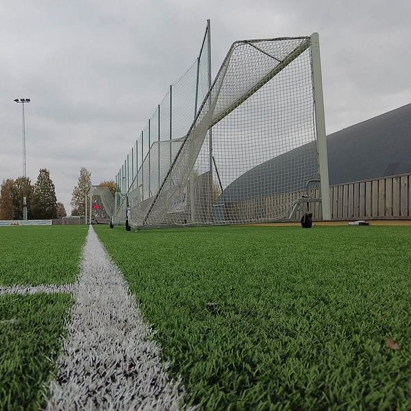 En delad bild som visar två sektioner: till vänster en man i uniform som ger en intervju och till höger en grön konstgräsmatta med en fotbollsmålvakt och mål i bakgrunden under en mulen himmel.