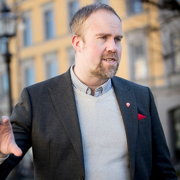 En man med kort hår och skägg. Klädd i skjorta, en ljus tröja och en mörk kavaj.