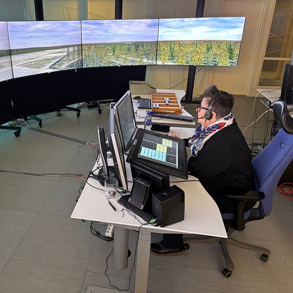 En person i arbetskläder sitter vid en datorstation med flera stora skärmar i en kontorsmiljö. Skärmarna visar en panoramautsikt över en flygplats som omges av skog och ett öppet landskap. Personen använder ett headset och ser koncentrerad ut medan de arbetar.