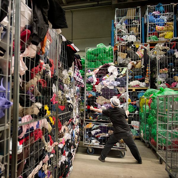 En person arbetar i en stor lagerlokal full med kläder som är sorterade i stora korgar. Korgarna står staplade på varandra och är fyllda med olika textilier i en mängd färger och mönster. Den anställde sträcker sig för att plocka upp eller sortera kläder från en av korgarna.