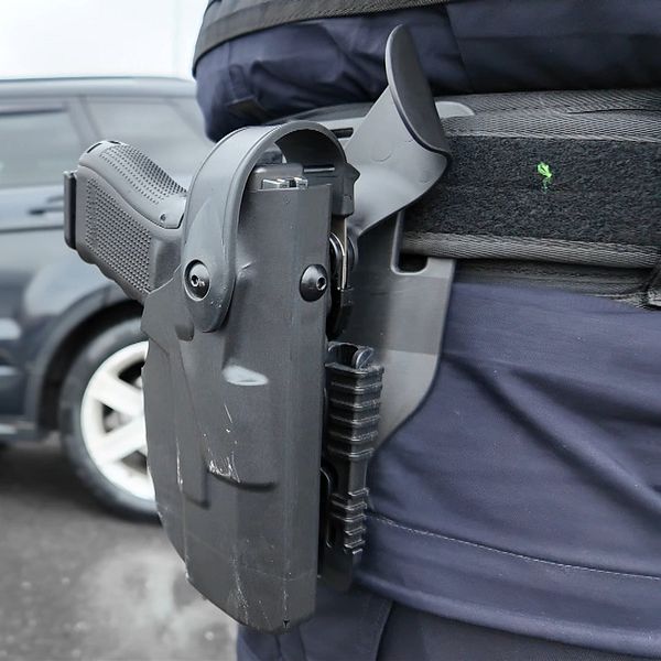 En tulltjänsteman står vid en bil och pratar med föraren, medan en annan person är synlig i förgrunden med en pistol i hölster. Scenen visar en ordningskontroll på en väg.