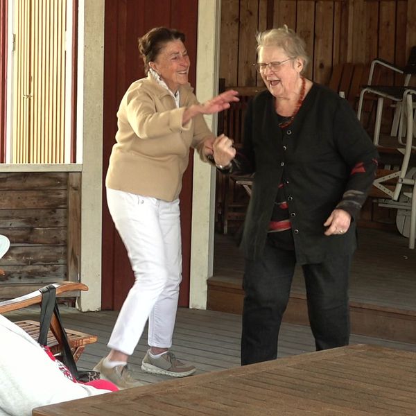 Två äldre kvinnor dansar på något som kan vara en dansbana, I förgrunden ser man en person i rullstol med ett hatt på huvudet och ytterligare ett huvud bakifrån (kvinna, kortklippt blont hår, glasögon).