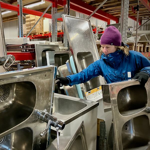 En person står bland en stapel av rostfria handfat i ett lager. Handfaten är i olika storlekar och former, och omgivningen visar reoler och annan verktygsmateriel. Belysningen är inomhus med en industriell känsla.