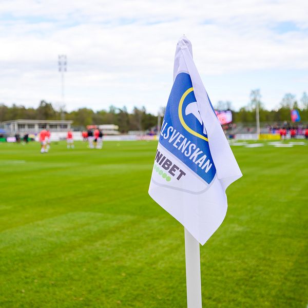 Hörnflagga med ”Allsvenskan” på fotbollsplan, spelare värmer upp i bakgrunden.