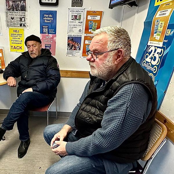 Tre äldre män sitter och pratar i ett rum med lottoaffischer på väggarna.