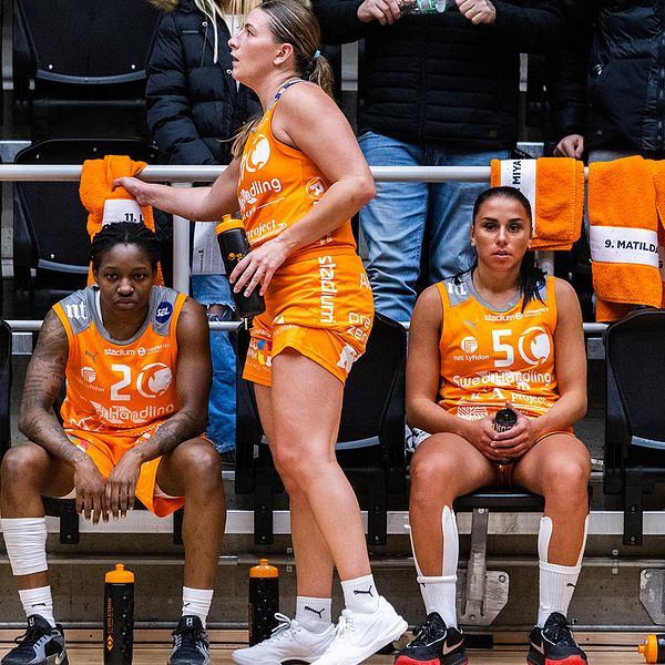 En delad bild som visar två scener från en basketballmatch. Den vänstra sektionen visar en spelare i en vit tröja med nummer 10, som är i rörelse på planen. Den högra sektionen visar två spelare, en i en orange tröja med nummer 20 och en annan i en orange tröja med nummer 50, som sitter på bänken. En av dem verkar ge vägledning eller uppmuntran. I bakgrunden syns lagets färger och supportersaker.