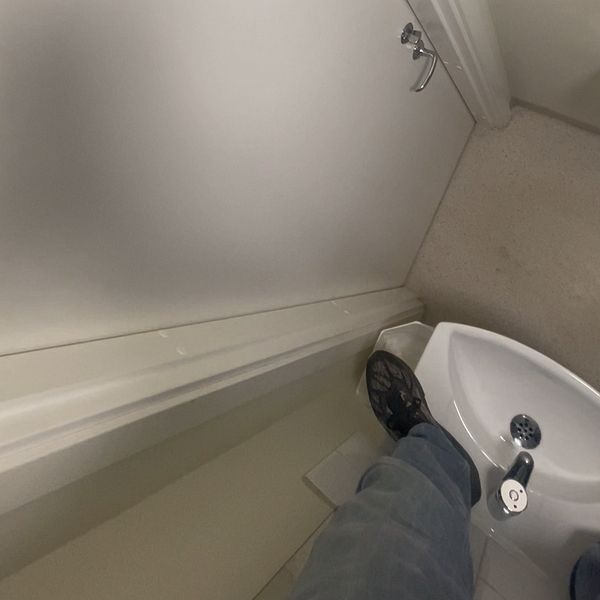 Delad bild. Vänstra sektionen visar en dörr i ett trångt badrum med en toalettpappershållare på väggen. Högra sektionen visar en persons fot på en liten tvättställsida, vilket antyder begränsat utrymme i badrummet.