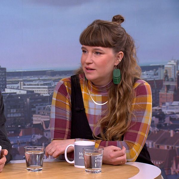 Två personer sitter vid ett bord i en tv-studio med stadsvy i bakgrunden.