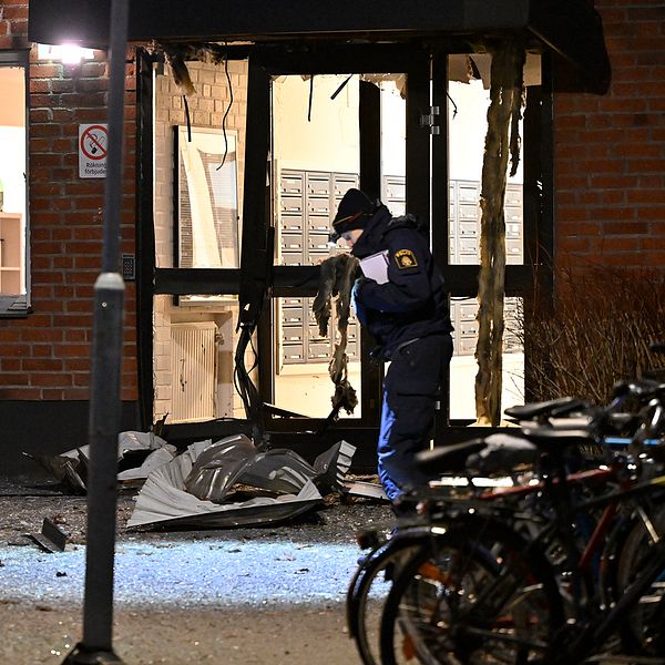 Jimmt Kirvesmäki står iförd mössa och jacka med mikrofon framför huset där det sprängdes. Till höger en polis framför den sprängda porten.
