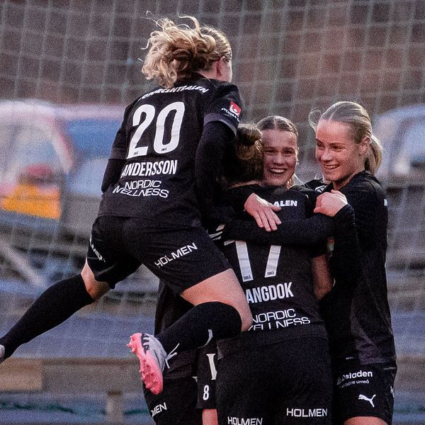 En grupp kvinnliga fotbollsspelare i svarta tröjor firar ett mål genom att omfamna varandra, medan en spelare i vit tröja observerar från sidan. Bakgrunden visar en fotbollsplan med gräs och en samtida infrastruktur.