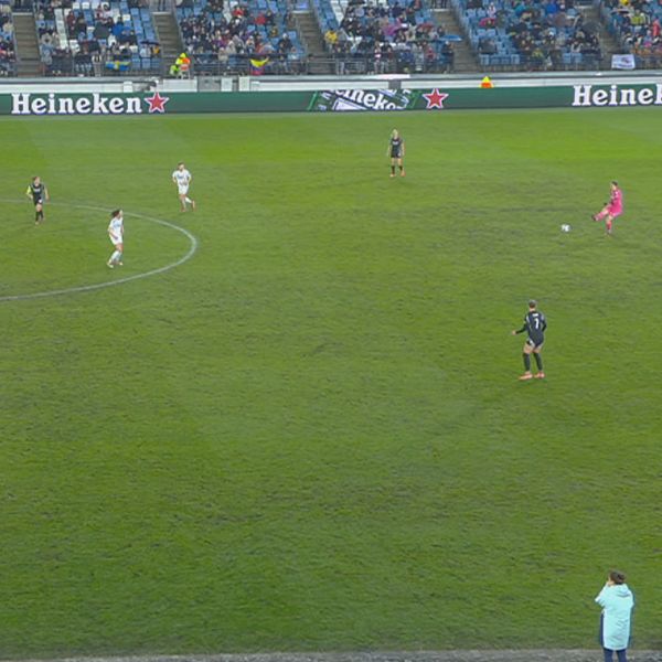 Fotbollsmatch på plan med spelare från två lag, åskådare i bakgrunden.