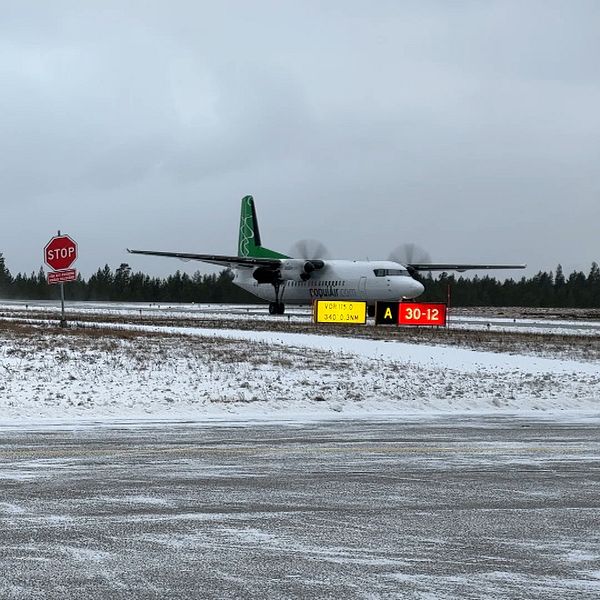 Ett lastplan med grön och svart färg är på väg att landa på en snöbelagd landningsbana. I förgrunden finns en röd stoppskylt och en vägskylt med information om bananummer 30-12. Området är omgiven av barrskog under en grå, molnig himmel.