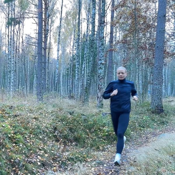En person joggar längs en stig i en skog med höga träd och höstfärgade löv. Naturen är lugn och omgivningen visar tydliga tecken på höst, med fjärran trädkronor och en grönskande mark.