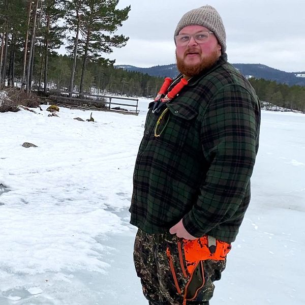 En man i mössa står utomhus på en frusen sjö bredvid ett hål i isen.