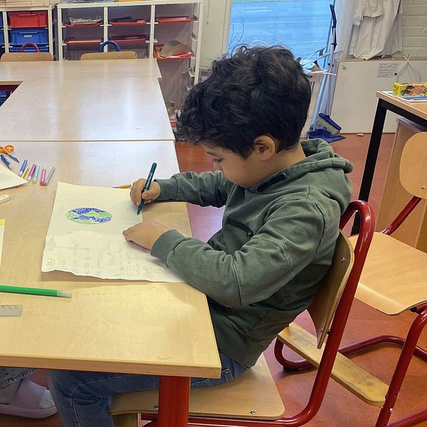 Tre barn sitter vid ett skolbord och ritar på papper med färgpennor. I bakgrunden finns hyllor för förvaring och klassrumsmaterial.