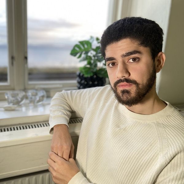 Jemal Shibli sitter i ett rum med en stor fönsteröppning som ger utsikt över en landskap i bakgrunden. Fönstret släpper in naturligt ljus, och en växt står på fönsterbrädan tillsammans med några dekorativa glasföremål. Jemal bär en ljus tröja och lutar sig avslappnat mot fönsterbrädan.
