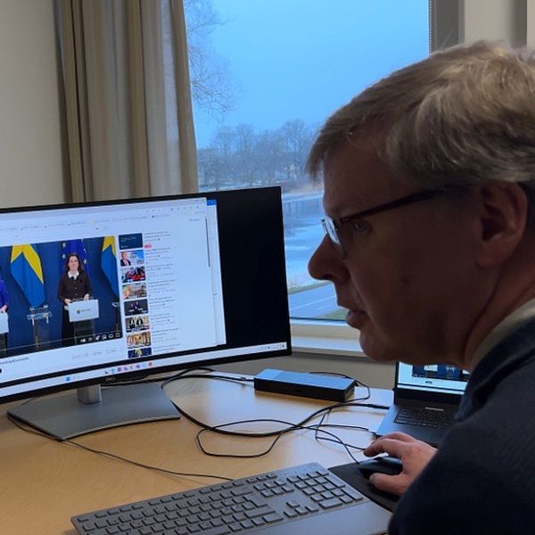 En person sitter vid en dator och tittar på en livestreamad presskonferens med svenska flaggor i bakgrunden. Skärmen visar två talare som står vid podium med mikrofoner under presentationen. I rummet syns även en teckning av byggnader på väggen och skrivmaterial på skrivbordet.