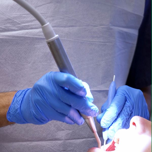 En tandläkare i blå skyddshandskar utför en behandling på en patient, med ett dentalverktyg riktat mot patientens mun. Bakgrunden visar en sjukvårdsinrättning med en tandläkarstol synlig.