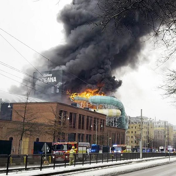 En delad bild som visar två sektioner. Den vänstra sektionen föreställer en man, berättar om, klädd i en blå skjorta med en krage. Den högra sektionen visar en stor byggnad, Lisebergs badhus Oceana, som brinner, med kraftig rök som stiger upp mot himlen, samt brandsläckningsfordon på plats.