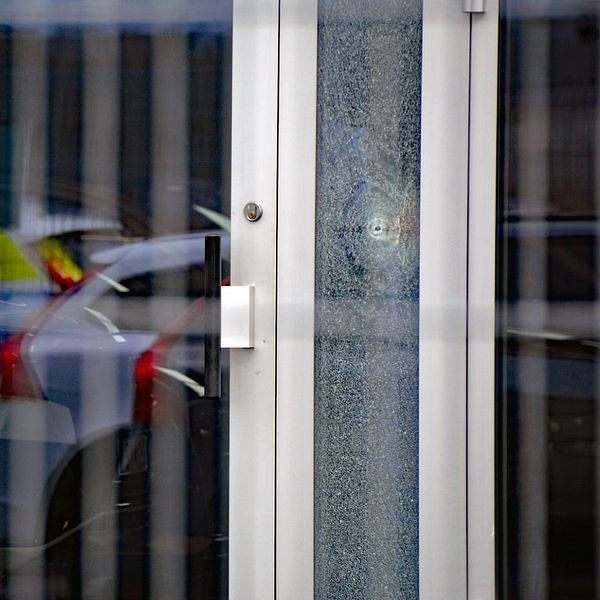 Två fösnterrutor med krossade glas och något som ser ut som ett skotthål. I fönstret speglas en polisbil.