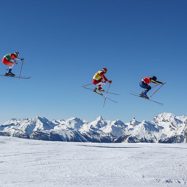 Fyra skidåkare hoppar i luften samtidigt under en skicross-tävling, med snötäckta berg i bakgrunden och klarblå himmel ovanför.