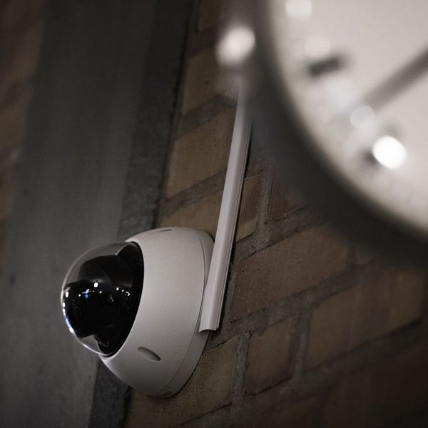 En delad bild där den första sektionen visar en person i mörka kläder sittande på en soffa, och den andra sektionen visar en övervakningskamera monterad på en tegelvägg, delvis täckt av en klocka.