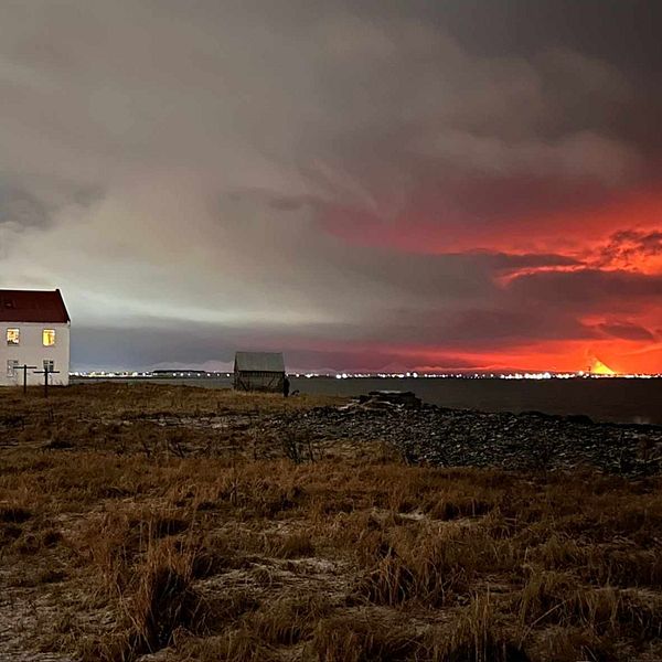 Kvinna med långt brunt hår som tittar in i kameran. Hus som står öde, till höger syns ett orange sken i molnen till följd av vulkanutbrott på Island.