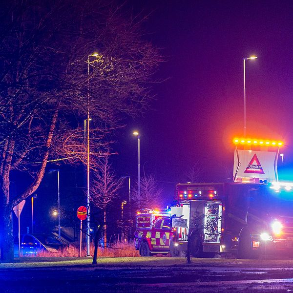 Utryckningsfordon med blinkande blåljus är uppställda längs en väg på natten. Ett fordon har även en skylt med varningsblinkers för att dirigera trafiken.