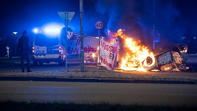 En banderoll med texten ”Nej till koranbränning. Missbruka ej yttrandefriheten” bredvid brinnande bil. Starka reaktioner i Rosengård i Malmö mot att koranen bränts av privatperson sommaren 2023.