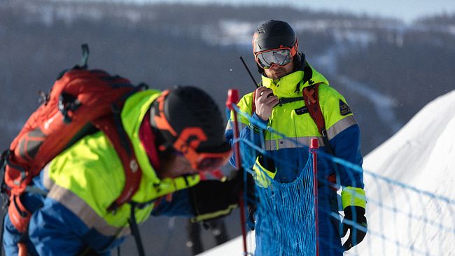 Två personer i färgglada vinterkläder arbetar på en snötäckt plats. Den ena personen använder en walkie-talkie och står nära en säkerhetsstaket av nät, medan den andra personen är i färd med att installera eller kontrollera staketet. Området har klart väder med snöiga fjäll i bakgrunden.