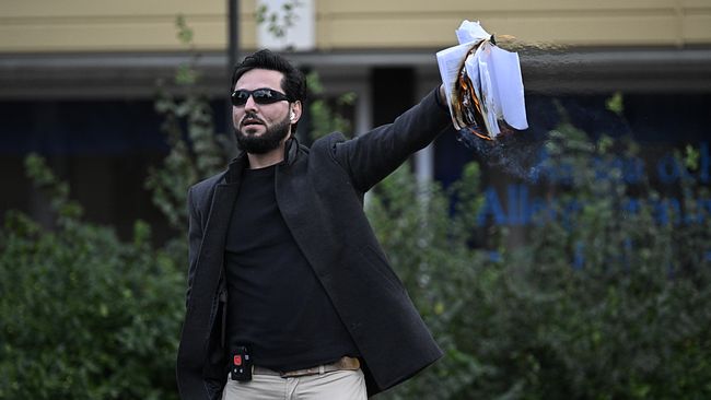 En person iklädd mörk jacka och ljusa byxor står med en bunt papper i ena handen. Papperen verkar delvis vara antända, och en rykande effekt syns från högen. Bakgrunden visar en buske och en byggnad.
