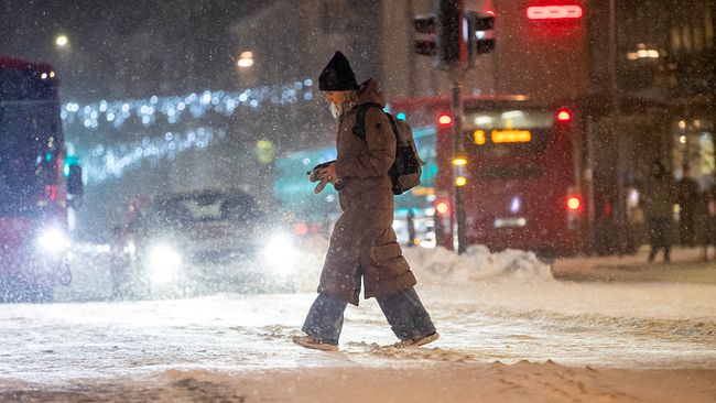 En person i vinterkläder går över en snötäckt gata. Snö faller och det finns billyktor och trafik i bakgrunden. Städ gatuskylten är synlig i ljuset från bilarna.