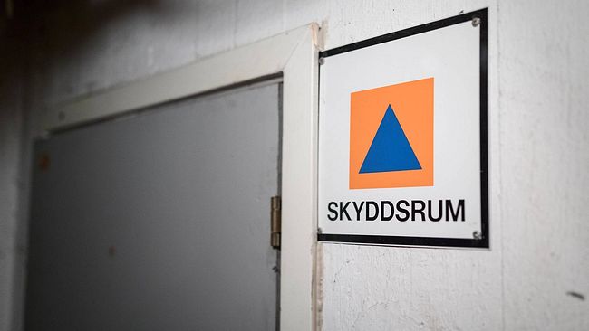 Bild på en kvadratisk skylt med texten ”skyddsrum”. På skylten finns en orange kvadrat med en blå triangel i.