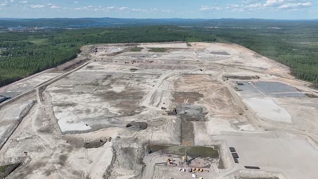 En panoramavy av ett stort område som visar en omfattande grävning och markavverkning. Landskapet är avskalat med flera stora maskiner synliga i området. Området omges av grönska och har en klar himmel i bakgrunden.
