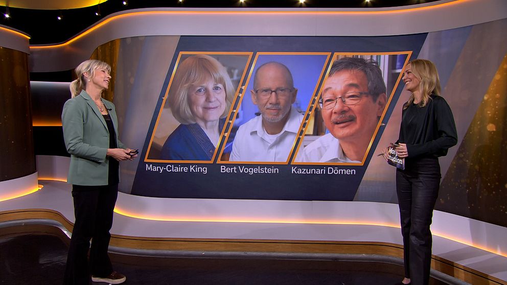 Två presentatörer står framför en stor skärm som visar tre sektioner med bilder av personer, markerade med namn: Mary-Claire King, Bert Vogelstein och Kazunari Dōmen. Presentatören till vänster har på sig en grön kavaj, medan den till höger bär en svart outfit.