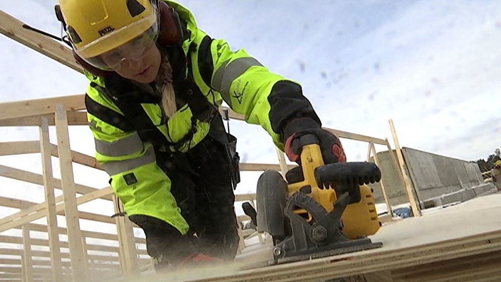 Kvinna iklädd varselkläder och hjälm sågar planka på jörnträhus i Jörn.