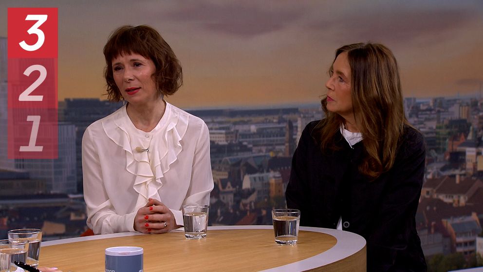 Två personer sitter vid ett runt bord i en tv-studio. De har varsitt glas vatten framför sig. Bakom dem är en bakgrund som föreställer en stadsvy.
