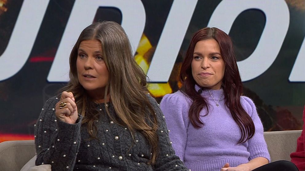 Två kvinnor sitter i en tv-studio under en diskussion. Den första kvinnan i grå tröja med pärlor gestikulerar medan den andra i lila tröja sitter med armarna i knät, vilket tyder på en engagerad dialog. Bakgrunden visar en suddig, färgglad grafik som antyder ett samtalsämne av intresse.
