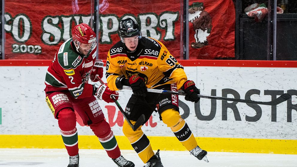 Två ishockeyspelare är i en fartfylld situation på isen, där den ena spelaren i röd tröja (nummer 2) försöker bryta puckkontakten med den andra spelaren i gul tröja (nummer 89). Arenan är fylld med fans och en stor banner syns i bakgrunden.