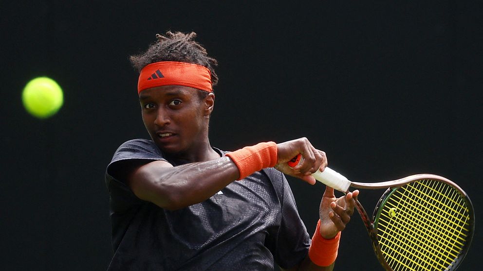 En tennisspelare i en svart tröja och med en orange svettband spelar en boll som syns i luften. Spelaren klipper en backhand med sin racket, med fokus på motståndet. Bakgrunden är mörk för att framhäva aktionen.
