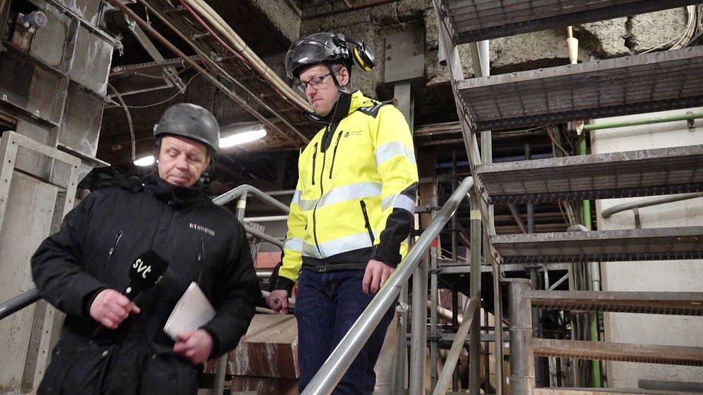 Två personer iklädda skyddskläder och hjälmar befinner sig i en industriell miljö på en trappa. Den ena bär en gul säkerhetsjacka och den andra en svart jacka märkt med ”SVT”.