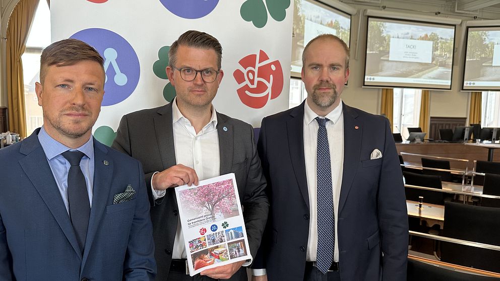 Tre män i kostym i ett sammanträdesrum, framför en skärm där partisymbolerna för moderaterna, socialdemokraterna och centerpartiet syns.